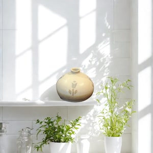 Puede incluir: Un jarrón de cerámica beige con un diseño de tulipán se exhibe en una estantería blanca. El jarrón tiene forma redondeada y una pequeña abertura en la parte superior. Dos pequeñas plantas en maceta con follaje verde están en primer plano.