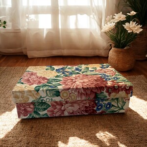 May include: A rectangular storage box featuring a vibrant floral pattern with large pink, yellow, and blue flowers set against a cream background. The box has a matching lid that covers the top, displaying a continuous botanical print of petals and leaves.