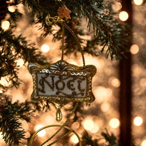 May include: A rectangular hanging ornament featuring the word NOEL embroidered in gold thread on a white fabric background, framed by gold sequins and metallic trim, suspended from a gold wire stand with a circular base and a small star topper.