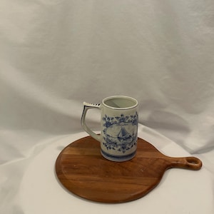 May include: A white ceramic mug with a blue sailboat design and handle. The mug is decorated with blue floral patterns and sits on a round wooden cutting board. The mug is approximately 10 cm tall.