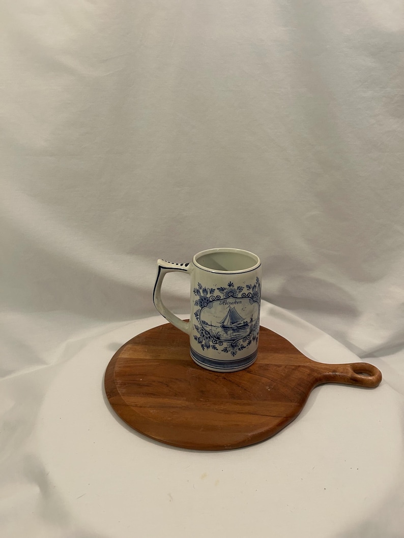 May include: A white ceramic mug with a blue sailboat design and handle. The mug is decorated with blue floral patterns and sits on a round wooden cutting board. The mug is approximately 10 cm tall.