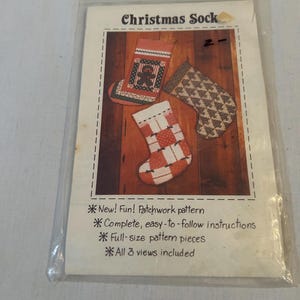 Peut inclure: Un patron de couture intitulé Christmas Sock présentant trois modèles de chaussettes en patchwork. Le paquet inclut des instructions, des pièces de patron en taille réelle et trois vues. La couverture montre un motif de bonhomme en pain d'épice et des motifs géométriques dans des tons rouge, blanc et marron.