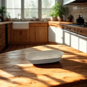May include: A speckled, oval-shaped ceramic serving dish sits on a wooden kitchen countertop. The dish is light gray with dark speckles. The kitchen features wooden cabinets, a white sink, and a window with natural light.