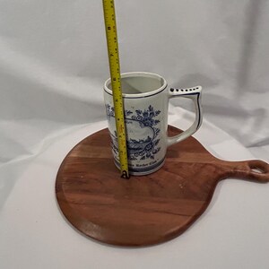 May include: A white ceramic mug with blue floral patterns and a handle, approximately 13 cm tall. The mug sits on a round wooden chopping board. A yellow measuring tape is next to the mug.