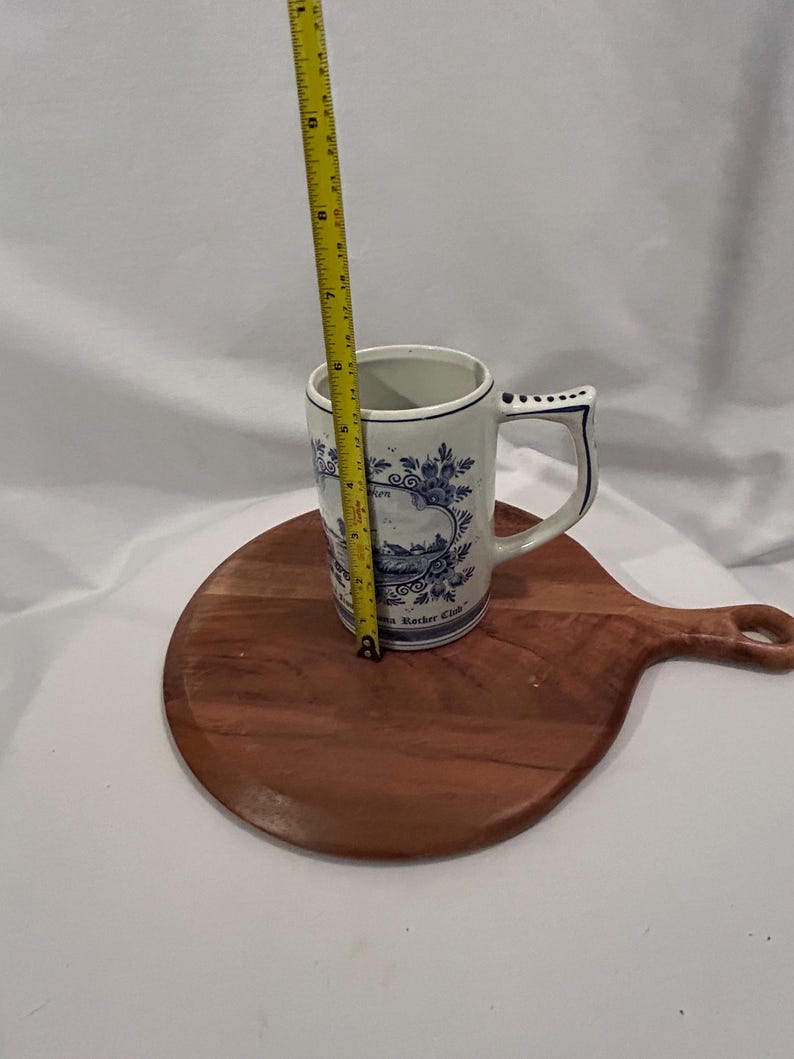 May include: A white ceramic mug with blue floral patterns and a handle, approximately 13 cm tall. The mug sits on a round wooden chopping board. A yellow measuring tape is next to the mug.