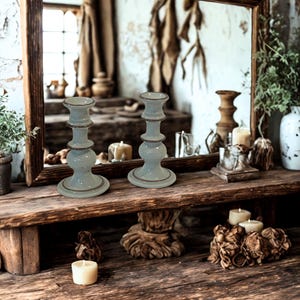 May include: Two light blue ceramic candle holders with a gold rim sit on a rustic wooden shelf. A large wooden framed mirror reflects the candle holders and other decorative items. The shelf also holds candles and other decor.