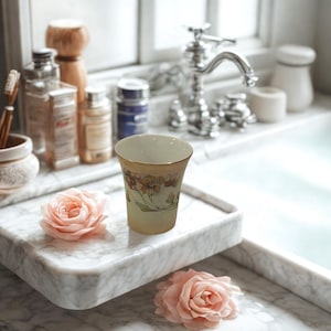 Pode incluir: Um copo de estilo vintage, amarelo pálido com um design floral e acabamento dourado, está sobre uma bandeja de mármore. Duas rosas cor-de-rosa são colocadas na bandeja. O fundo inclui uma pia, uma torneira e vários artigos de higiene pessoal.
