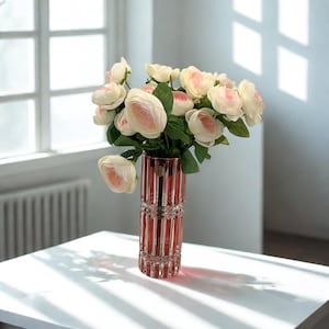 May include: A red crystal vase filled with light pink and white roses sits on a white table. The vase has a vertical ribbed design. The flowers have green leaves. Sunlight streams through a window in the background.