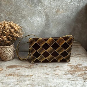 Peut inclure: Un sac à main rectangulaire avec un motif de losanges marron et beige. Le sac a une longue et fine bandoulière et une fermeture éclair. Le sac est fait d'une matière texturée et repose sur une surface en bois.