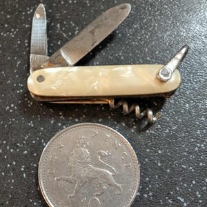 Pode incluir: Uma canivete vintage com cabo creme, uma pequena lâmina, um saca-rolhas e uma segunda lâmina. Uma moeda de 10 pence está ao lado da faca. A faca tem aproximadamente 7.6 cm de comprimento.