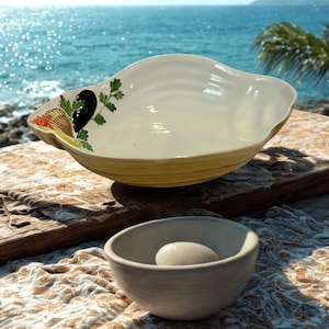 May include: A large, off-white ceramic serving bowl with a scalloped edge, decorated with a painted design of olives, parsley, and a tomato. A smaller, gray ceramic bowl sits in front of it. The background shows the ocean.