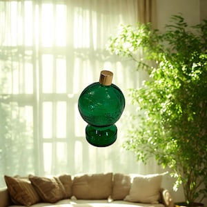 May include: A green glass decorative bottle with a gold-coloured stopper. The bottle has a round shape with a textured surface and sits on a small, round base. The bottle is displayed in a well-lit room with a sofa and a large plant.