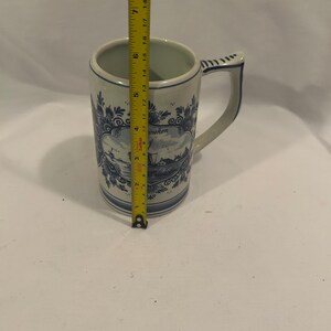 May include: A tall, white ceramic mug with a blue and white Dutch design. The mug has a handle and a rectangular panel with a landscape scene. The mug is approximately 7 inches tall.