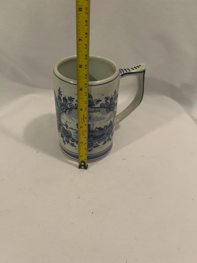 May include: A tall, white ceramic mug with a blue and white Dutch design. The mug has a handle and a rectangular panel with a landscape scene. The mug is approximately 7 inches tall.