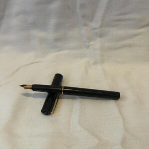May include: A black fountain pen with a gold nib and gold accents. The pen is lying on a white surface, with the cap placed next to the pen. The pen is a classic writing instrument.