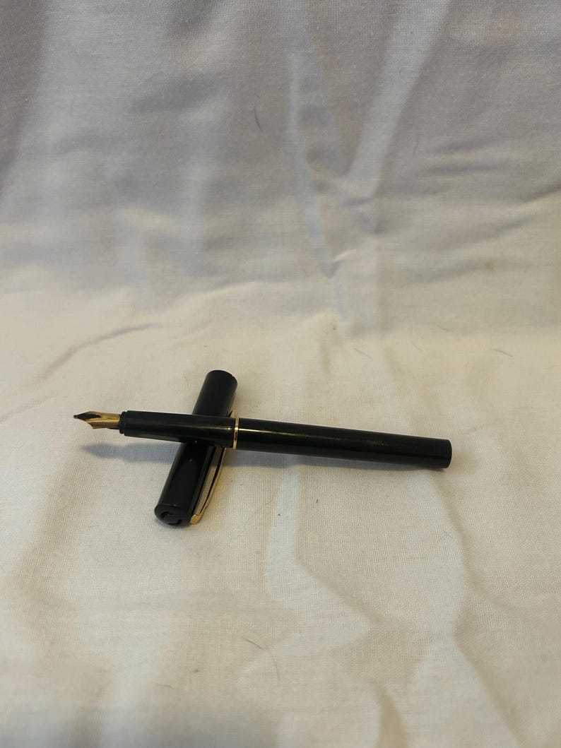 May include: A black fountain pen with a gold nib and gold accents. The pen is lying on a white surface, with the cap placed next to the pen. The pen is a classic writing instrument.