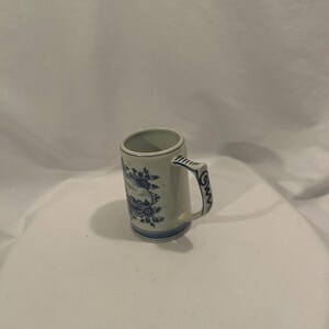May include: A small, off-white ceramic mug with a handle. The mug features a blue floral and landscape design around the body and handle. The rim and base are also trimmed with blue. The mug is approximately 4 inches tall.
