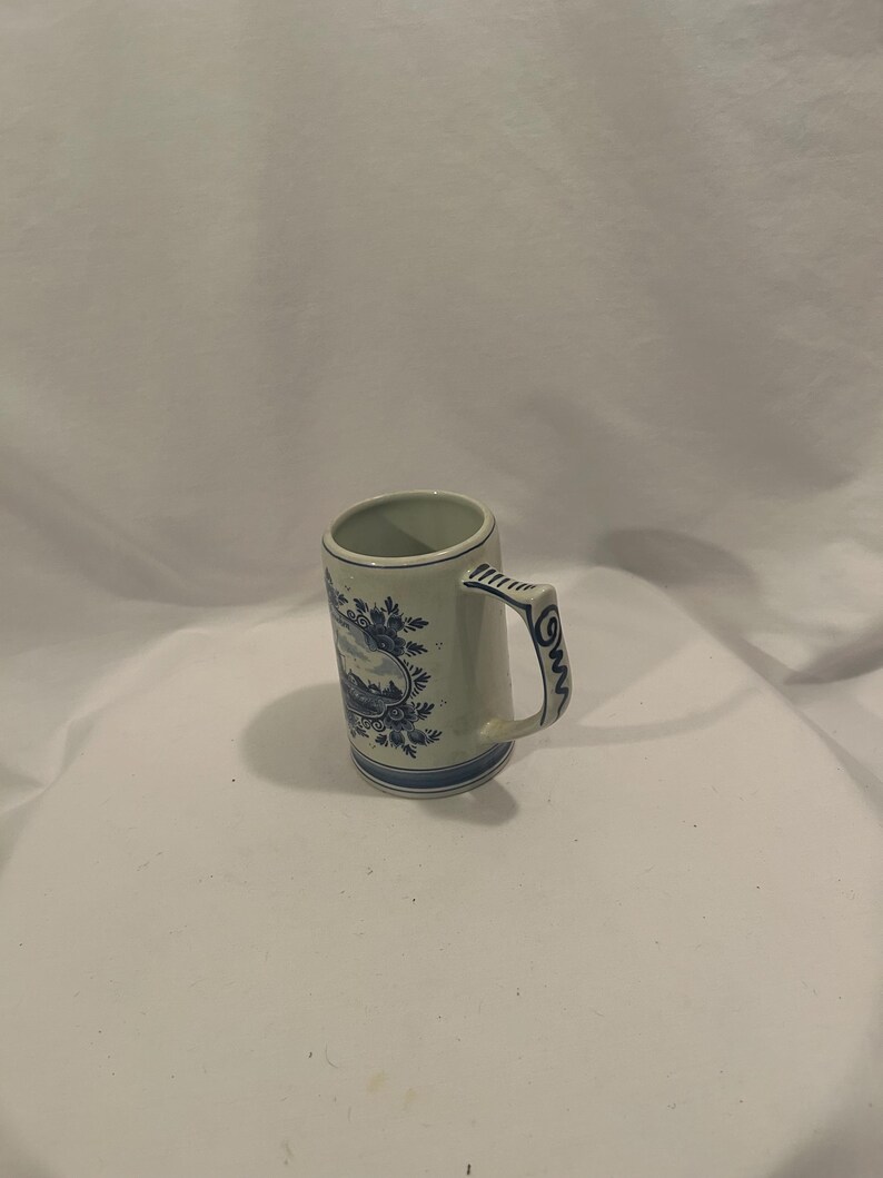 May include: A small, off-white ceramic mug with a handle. The mug features a blue floral and landscape design around the body and handle. The rim and base are also trimmed with blue. The mug is approximately 4 inches tall.