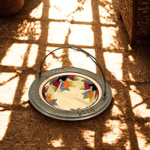 May include: A decorative serving dish with a metal rim and handle. The dish features a cream-colored interior with a floral pattern in shades of orange, blue, and purple. The dish is sitting on a brown woven surface.