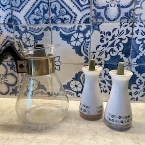 以下が含まれることがあります： 黒いハンドルと金色のアクセントが付いた透明なガラスのコーヒーポットと、花の模様が入った白の塩コショウ入れが2つ並んでいます。背景には青と白の模様のタイルがあります。