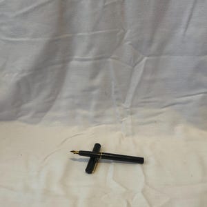 May include: A black fountain pen with a gold-colored nib and accents. The pen is lying on a white surface. The cap is open, revealing the nib. The pen is a writing instrument.