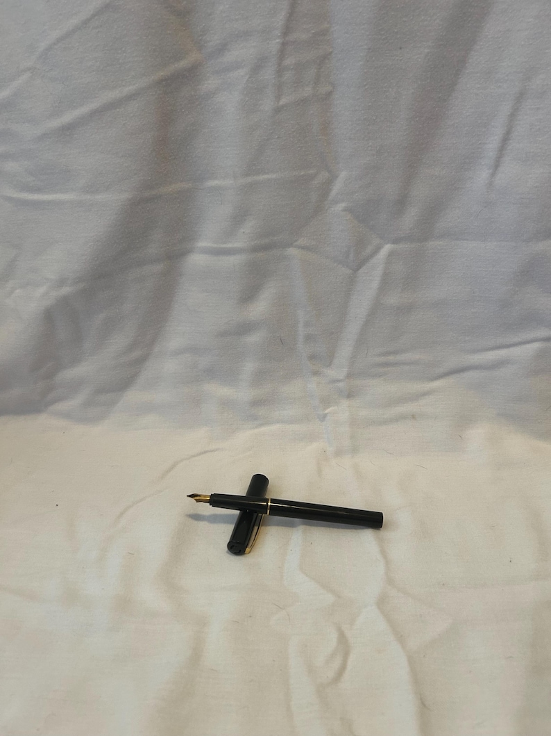 May include: A black fountain pen with a gold-colored nib and accents. The pen is lying on a white surface. The cap is open, revealing the nib. The pen is a writing instrument.