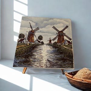 May include: A square ceramic tile painting depicting a Dutch landscape with three brown windmills, white houses, and a central canal reflecting the sky. The artwork features green grassy banks, a small fishing net in the water, and a cloudy sky.