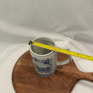 May include: A white ceramic mug with blue floral and landscape designs. The mug has a handle and the text "Reineken" and "Reineken Roster Club". The mug is approximately 10 cm tall.