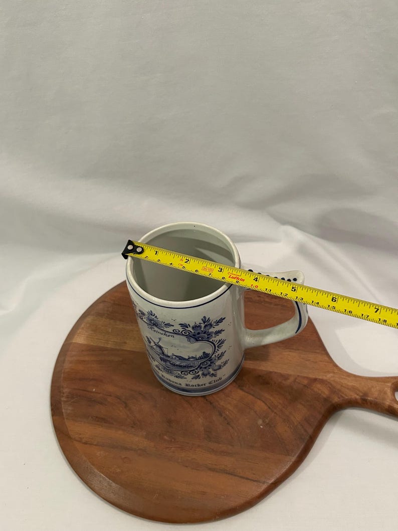 May include: A white ceramic mug with blue floral and landscape designs. The mug has a handle and the text "Reineken" and "Reineken Roster Club". The mug is approximately 10 cm tall.