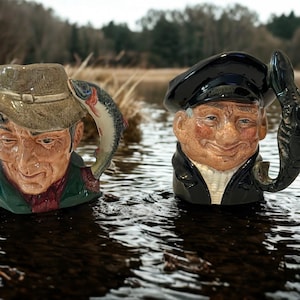 May include: Two ceramic mugs featuring elderly men, one with a hat and fish handle, the other with a cap and fish handle. The mugs are in water, with a blurred background of trees and water.