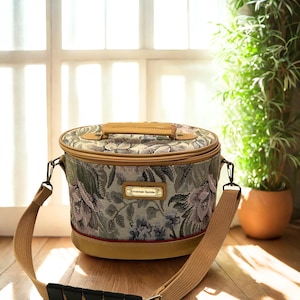 Pode incluir: Bolsa de viagem redonda com padrão floral em tons de verde, roxo e bege. Possui uma alça de ombro e uma alça bege, com acabamentos dourados e o texto "American Tourister" em uma pequena etiqueta.