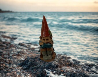 Könnte beinhalten: Eine kleine Gartenzwergfigur mit roter Zipfelmütze und blauer Jacke sitzt an einer felsigen Küste. Der Zwerg blickt nach unten, die Arme verschränkt. Der Hintergrund zeigt einen bewölkten Himmel und Meereswellen.