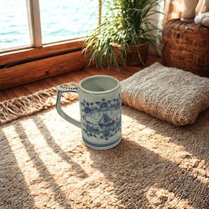May include: A ceramic mug with a blue and white nautical design featuring a sailboat. The mug has a handle with a decorative pattern and is set on a textured rug. The mug is cylindrical and has a light blue interior.