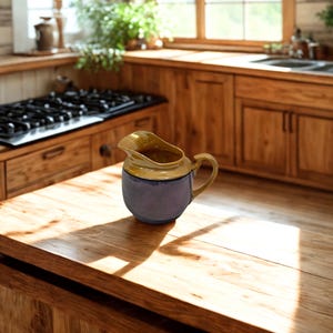 May include: A ceramic pitcher with a rounded body and a handle. The top is a light yellow color, and the body is a dark blue-purple. The pitcher is sitting on a wooden countertop in a kitchen setting. The kitchen has wooden cabinets and a gas stove.