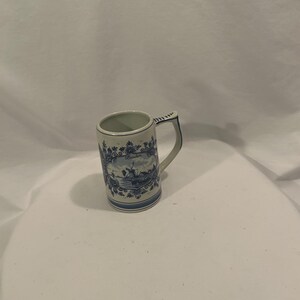 May include: A ceramic mug with a handle, featuring a blue and white design. The mug has a rectangular shape with a detailed illustration of a landscape scene. The handle is adorned with a blue pattern, and the rim has a decorative border.