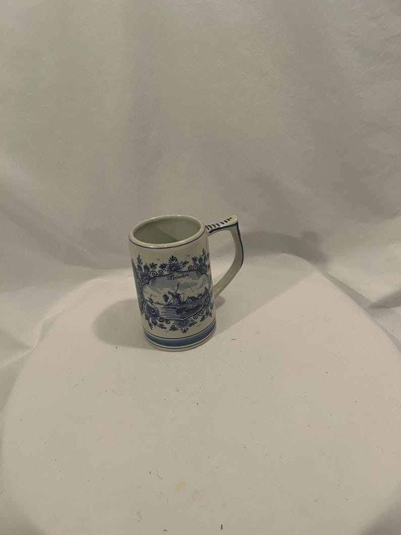 May include: A ceramic mug with a handle, featuring a blue and white design. The mug has a rectangular shape with a detailed illustration of a landscape scene. The handle is adorned with a blue pattern, and the rim has a decorative border.