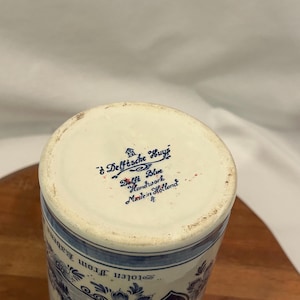 May include: A cylindrical Delft Blue ceramic vase with a floral design and the text "Delftsche Huyz Delft Blue Handwork Made in Holland". The vase has a white base and blue floral patterns.