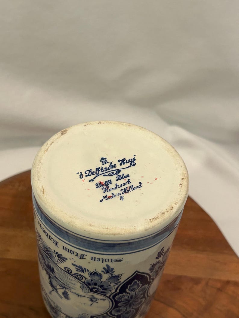 May include: A cylindrical Delft Blue ceramic vase with a floral design and the text "Delftsche Huyz Delft Blue Handwork Made in Holland". The vase has a white base and blue floral patterns.