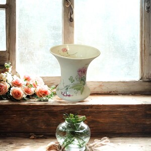 May include: A cream-colored ceramic vase with a flared rim and a painted floral design featuring pink flowers and green leaves, positioned above a small, clear glass spherical vase containing green stems and foliage.