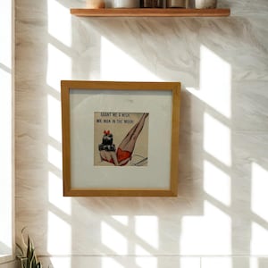 May include: A framed print with a retro illustration of a woman in red swimwear, with the text "Grant me a wish, Mr. Man in the Moon!" The print is in a light brown wooden frame, hanging on a white tiled wall. A wooden shelf with jars and plants is above the frame.