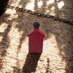 May include: A red fire alarm station replica with a gabled roof and a black spherical knob on top. The front surface features embossed text that reads FIRE ALARM and includes a small rectangular detail near the base of the red housing.