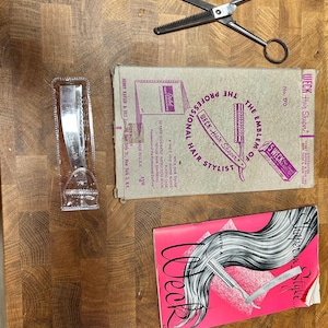 May include: A collection of vintage barber tools on a wooden surface. Includes shears, a clear plastic razor holder, and two boxes. One box reads "WECK Hair Shaper" and the other is a pink booklet with the word "Week".