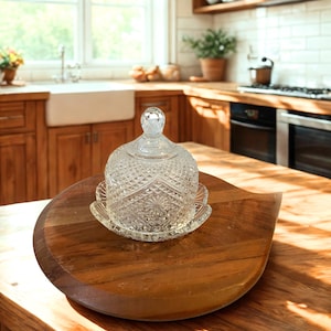 May include: A clear glass butter dish with a domed lid featuring a diamond-cut pattern and a small spherical knob handle, resting on a circular wooden board with a smooth finish and a distinct pointed edge on one side.