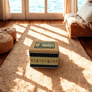 May include: A rectangular metal storage tin with a dark blue base featuring a decorative band of light green and gold patterns. The lid displays a painted illustration of a windmill near a body of water under a soft sky, framed by a gold border.
