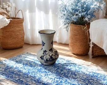 May include: A ceramic vase with a cream-colored base and blue floral patterns, featuring a central illustration of a windmill. The vase has a flared rim and a rounded body, sitting on a rectangular rug with a blue and white intricate pattern.