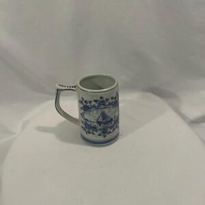 May include: A white ceramic mug with a blue floral and sailboat design. The mug has a handle and a cylindrical shape. The design includes a sailboat, flowers, and the word "Groningen".