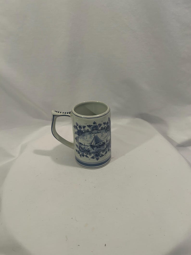 May include: A white ceramic mug with a blue floral and sailboat design. The mug has a handle and a cylindrical shape. The design includes a sailboat, flowers, and the word "Groningen".