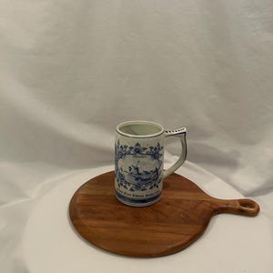 May include: A blue and white ceramic mug with a handle, decorated with a windmill scene and floral motifs. The mug sits on a round wooden chopping board. The mug has the text "Heineken" and "From Rabena Racker Club".