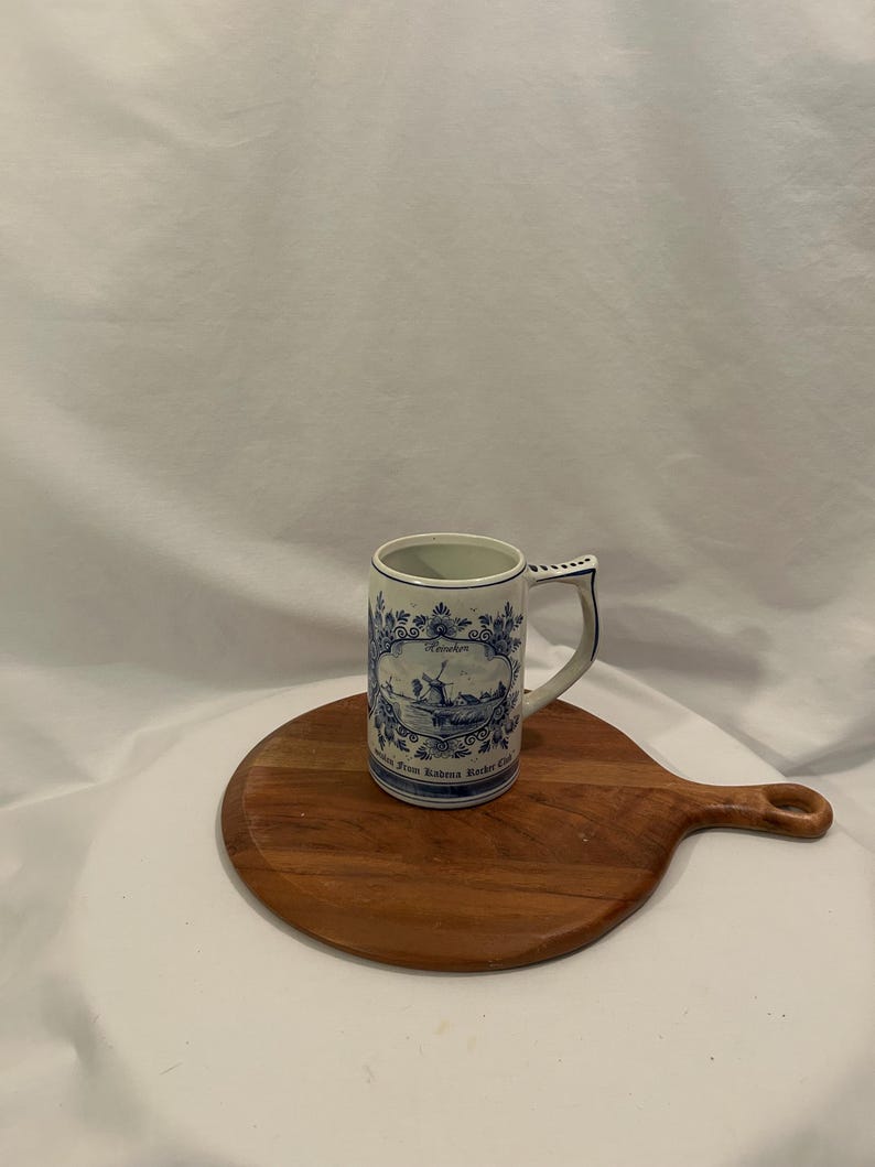 May include: A blue and white ceramic mug with a handle, decorated with a windmill scene and floral motifs. The mug sits on a round wooden chopping board. The mug has the text "Heineken" and "From Rabena Racker Club".