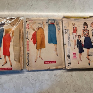 May include: Three vintage sewing pattern envelopes featuring illustrations of skirts, blouses, and dresses. The Simplicity and McCall's brand envelopes display various garment styles, including pencil skirts and shift dresses, with printed text and prices.